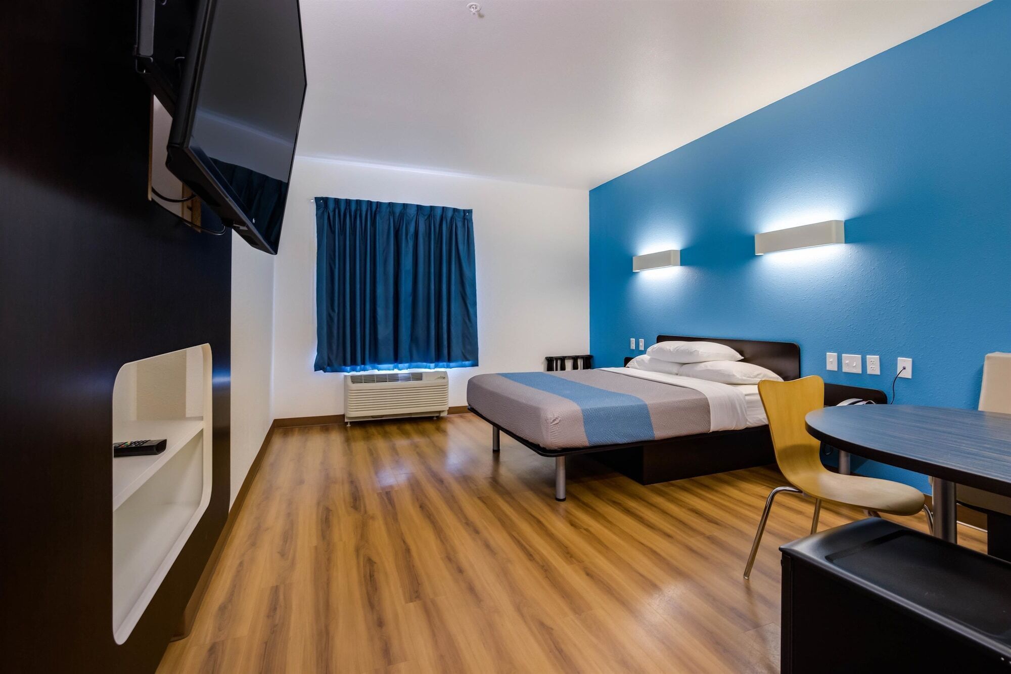Quarto de hotel com cama, escrivaninha, cadeira, televisão e janela com cortinas.