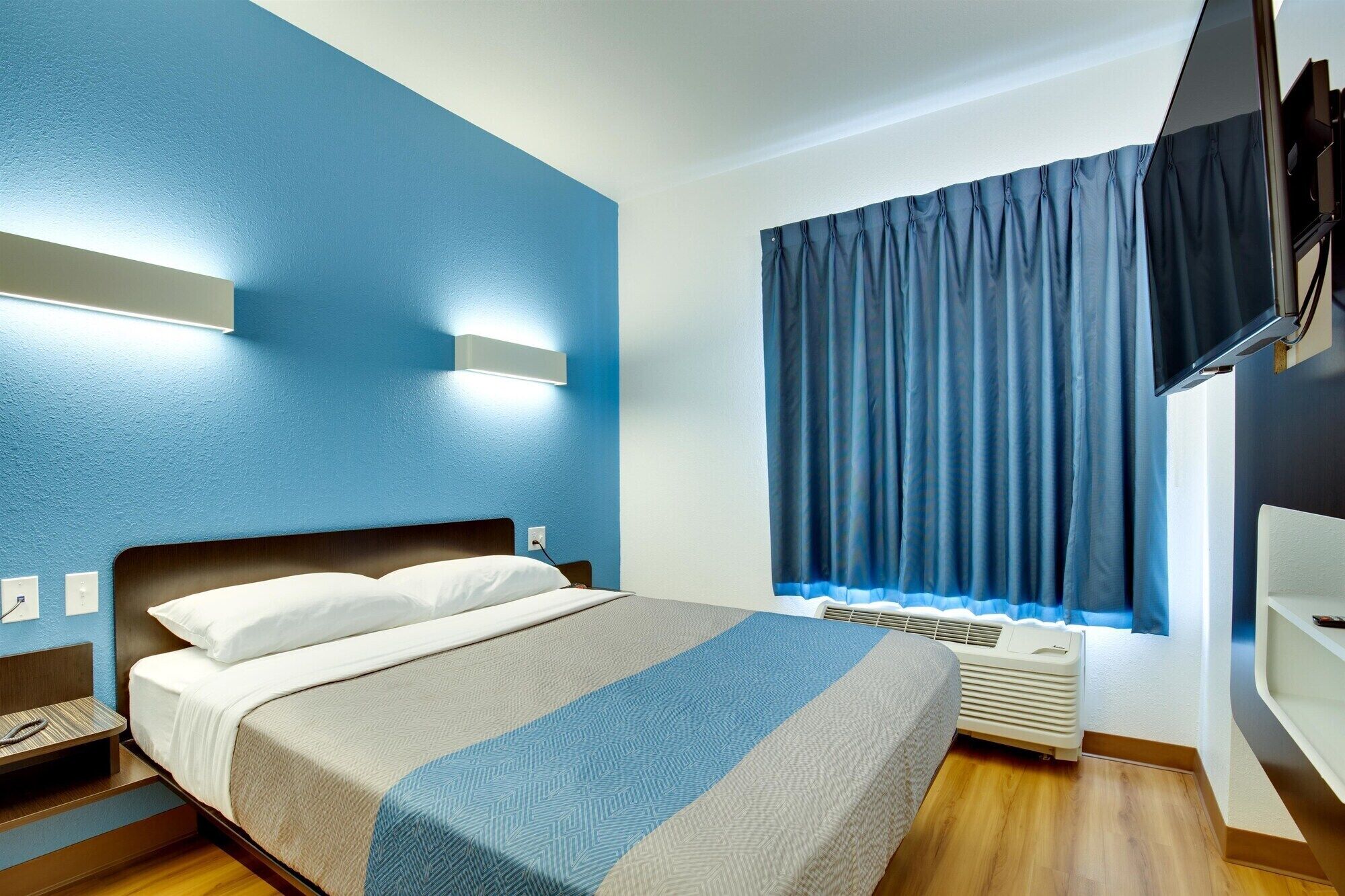 A modern hotel room with a bed, a wall-mounted TV, a window with curtains, and a built-in air conditioning unit.