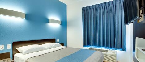 A modern hotel room with a bed, a wall-mounted TV, a window with curtains, and a built-in air conditioning unit.