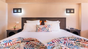 Un letto rifatto con cuscini bianchi e un cuscino con una vivace fantasia tropicale, affiancato da due comodini con lampade, in una stanza con una parete beige semplice e applique decorative.