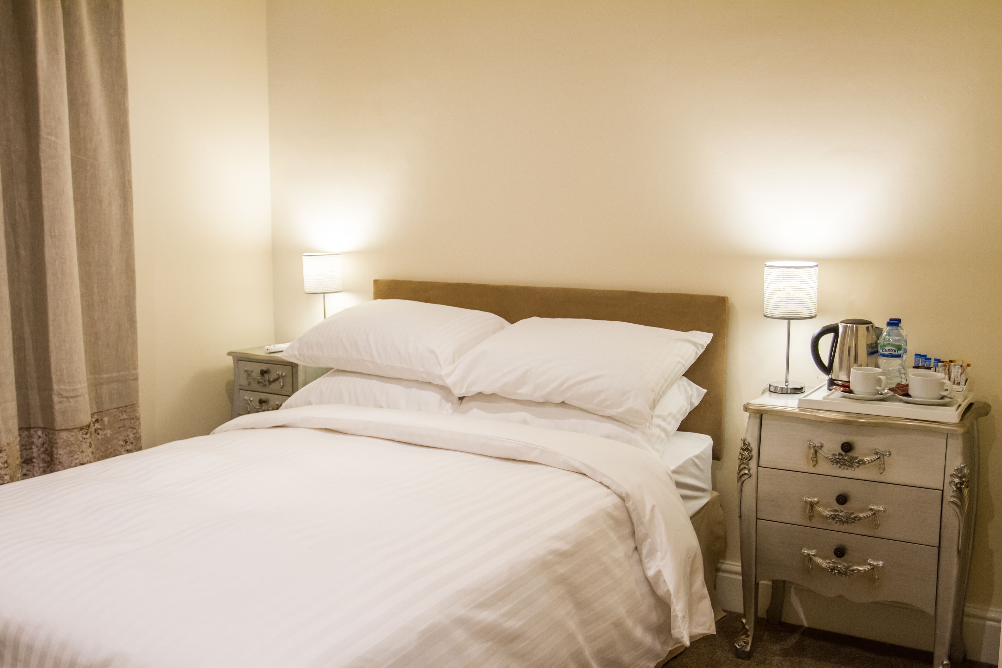 A neatly made bed with white linens, a bedside table with a lamp, and a small drawer unit.