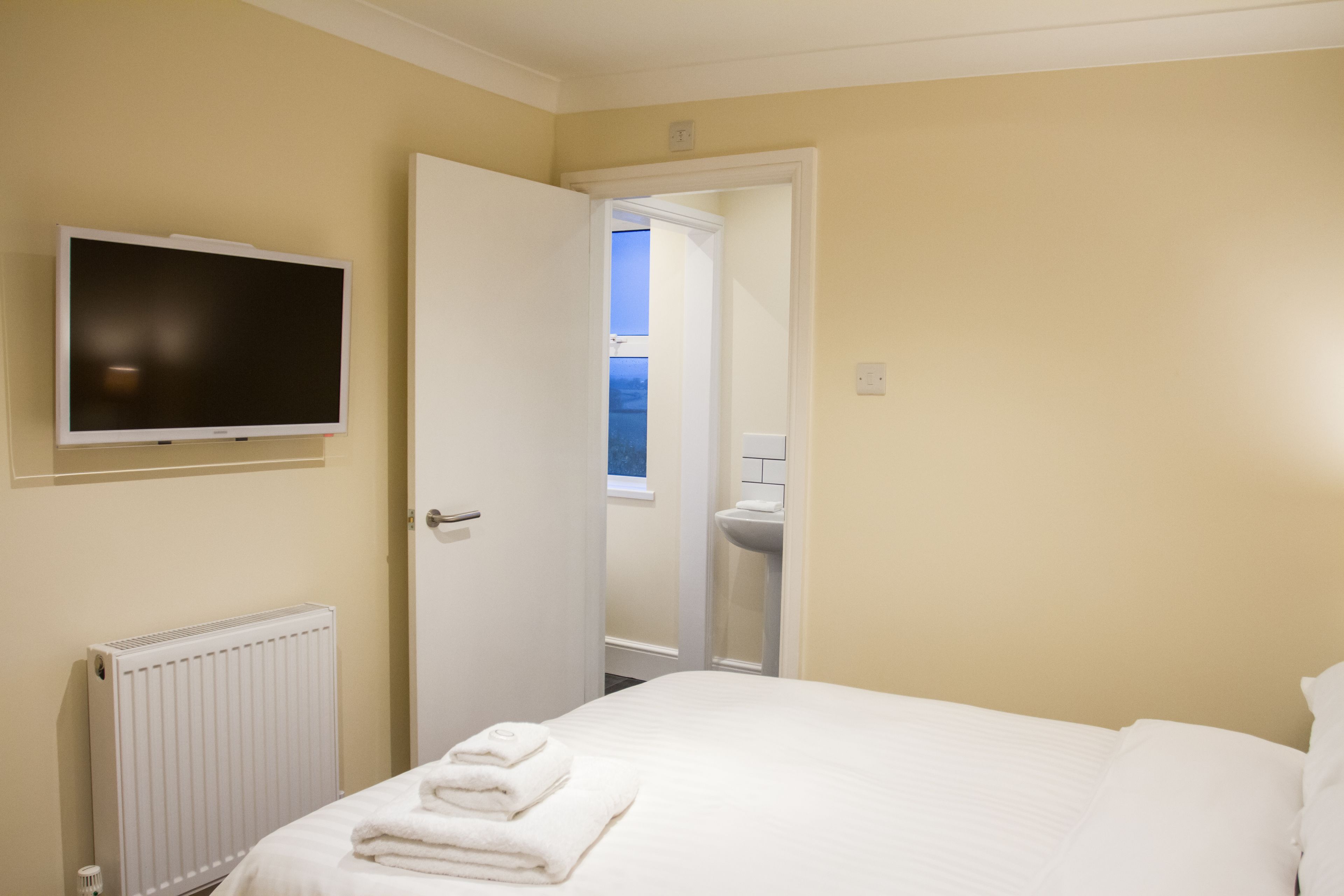 A hotel room with a bed, a TV mounted on the wall, a radiator, and a bathroom visible through an open door.