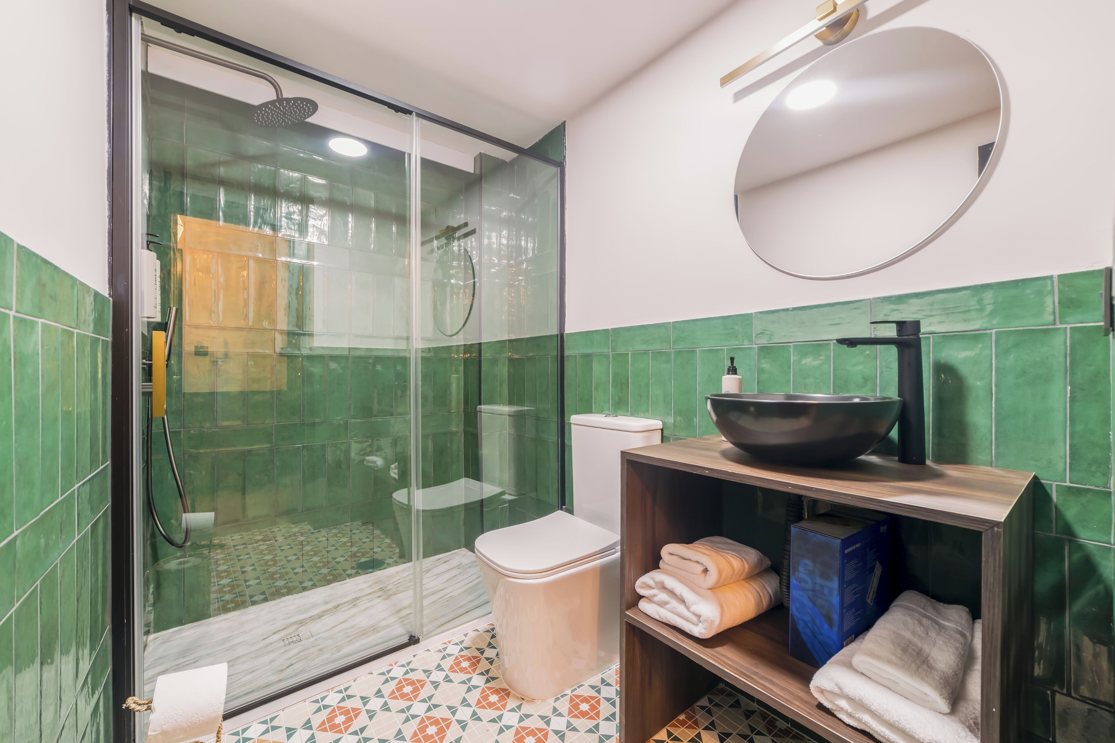 A bathroom with a glass-enclosed shower, a toilet, a sink with a bowl, and a shelf with towels and toiletries.