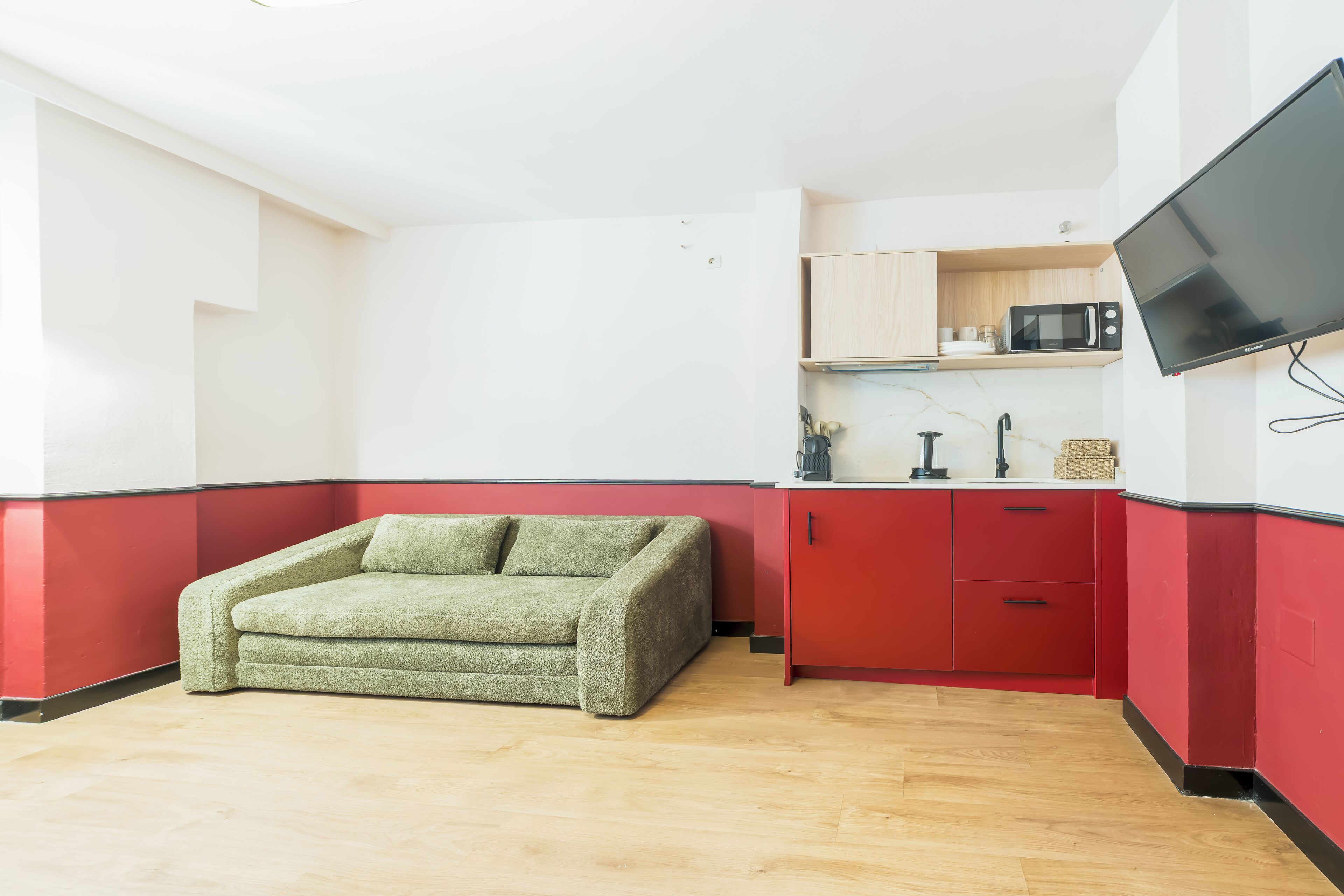 Un espace de vie compact avec un canapé vert, des meubles rouges, un four à micro-ondes et un téléviseur à écran plat fixé au mur.