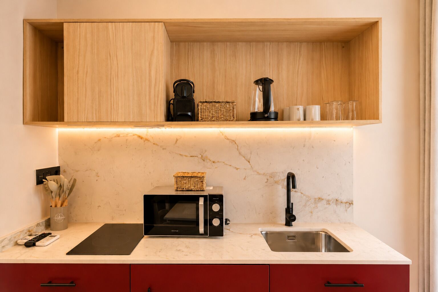 Cocina con microondas, tostadora y cafetera sobre una encimera de mármol, un lavamanos y un estante de madera encima.