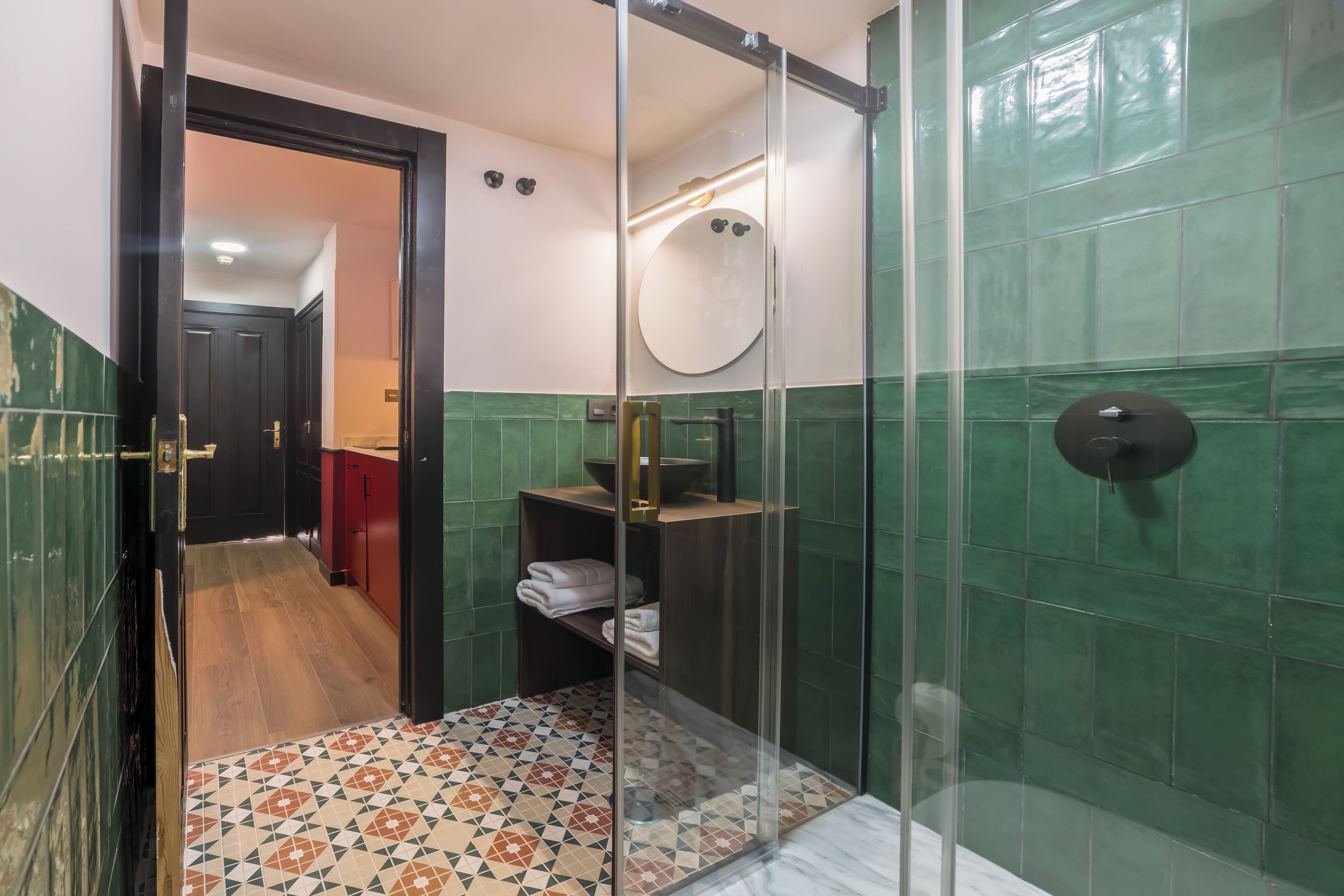 A modern bathroom with a glass shower enclosure, a wooden floor, and green tiled walls.
