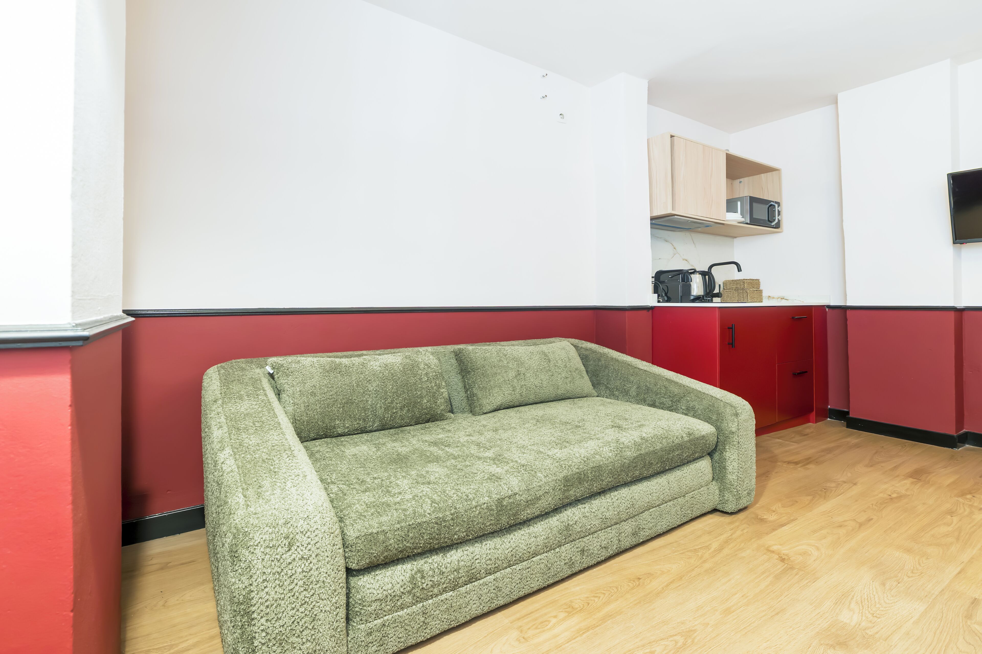 Una habitación con un sofá verde, suelo de madera, microondas y televisión en la pared.