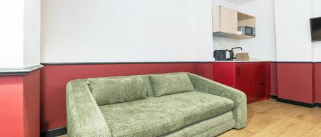 Una habitación con un sofá verde, suelo de madera, microondas y televisión en la pared.