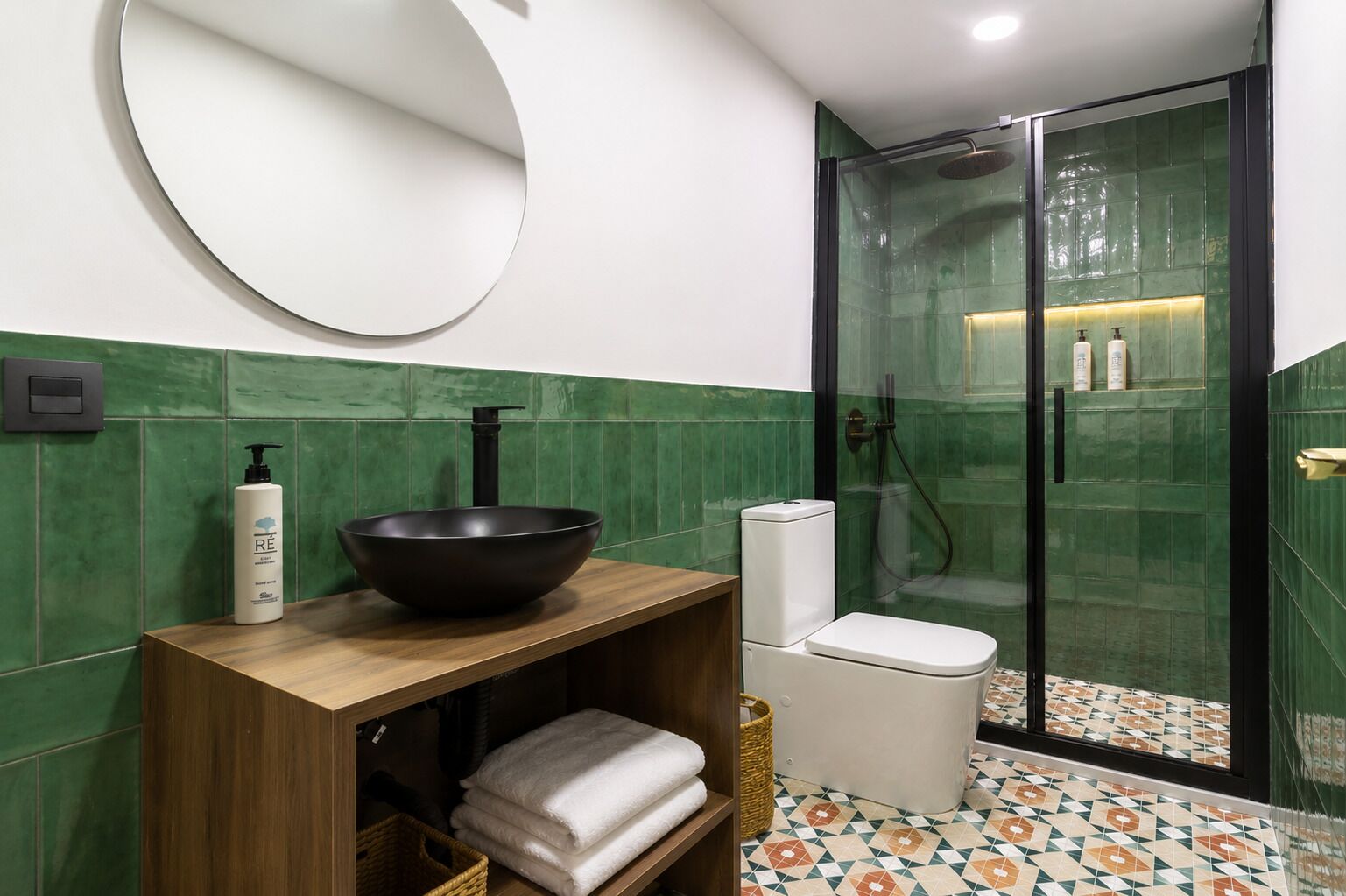 A bathroom with a wooden vanity, a black bowl sink, a toilet, a glass-enclosed shower, and a green tiled wall.
