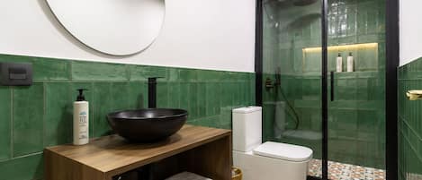 Un baño con tocador de madera, un lavabo de cerámica negra, inodoro, ducha con mampara de vidrio y pared con azulejos verdes.