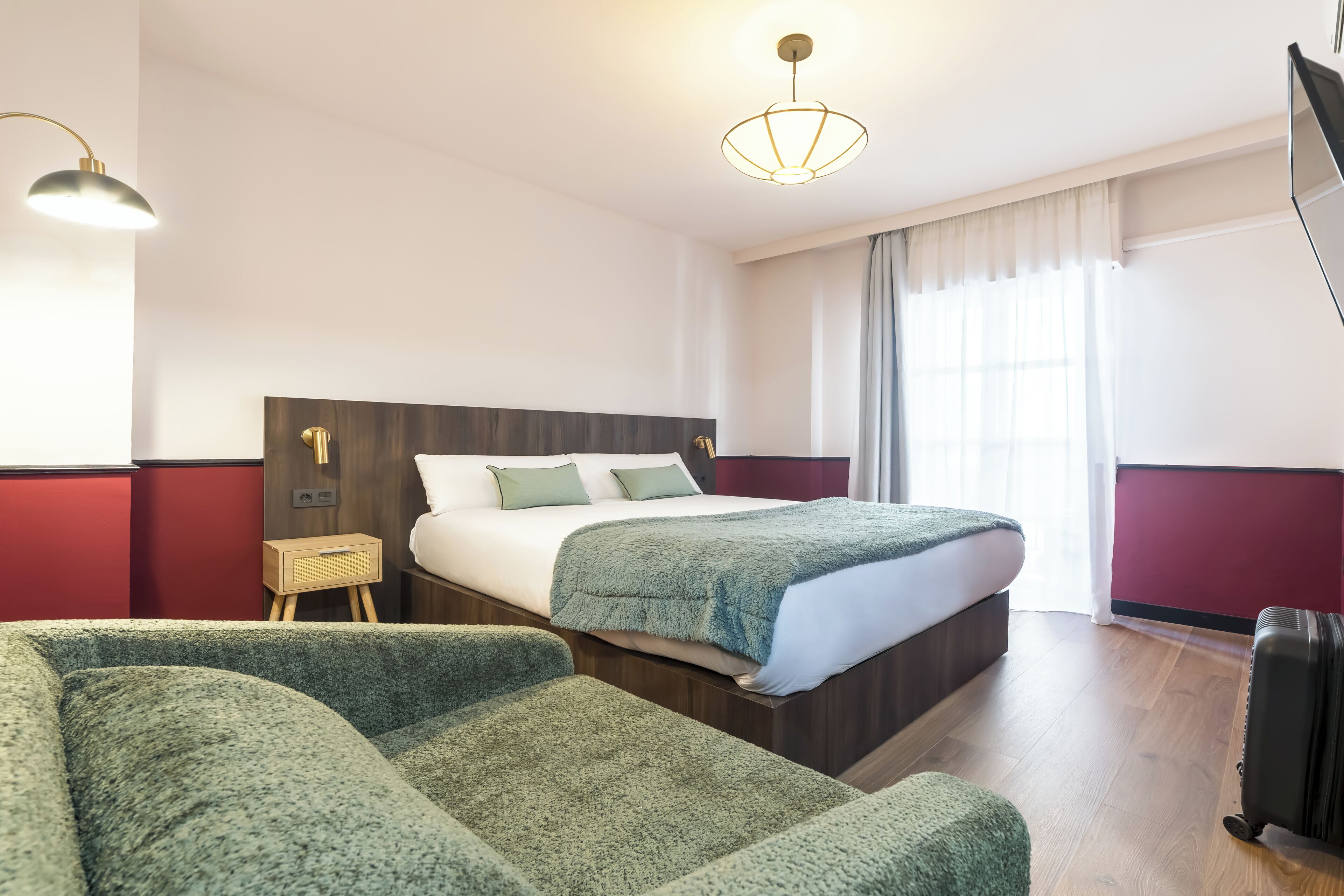 Een hotelkamer met een bed, twee groene banken, een nachtkastje, een lamp en een koffer.