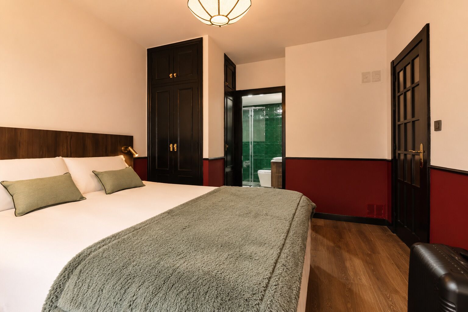 A hotel room with a bed, a bedside lamp, a wooden headboard, a red and white wall, and a door leading to a bathroom.