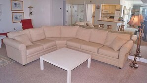 En stue med en beige hjørnesofa, et hvidt sofabord og et køkken i baggrunden.