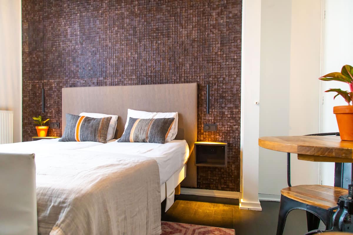 Une chambre d’hôtel moderne avec un mur d’accent orné d’un motif en briques, un lit bien fait avec deux oreillers, une petite table de chevet avec une lampe et une plante en pot posée sur une table en bois.