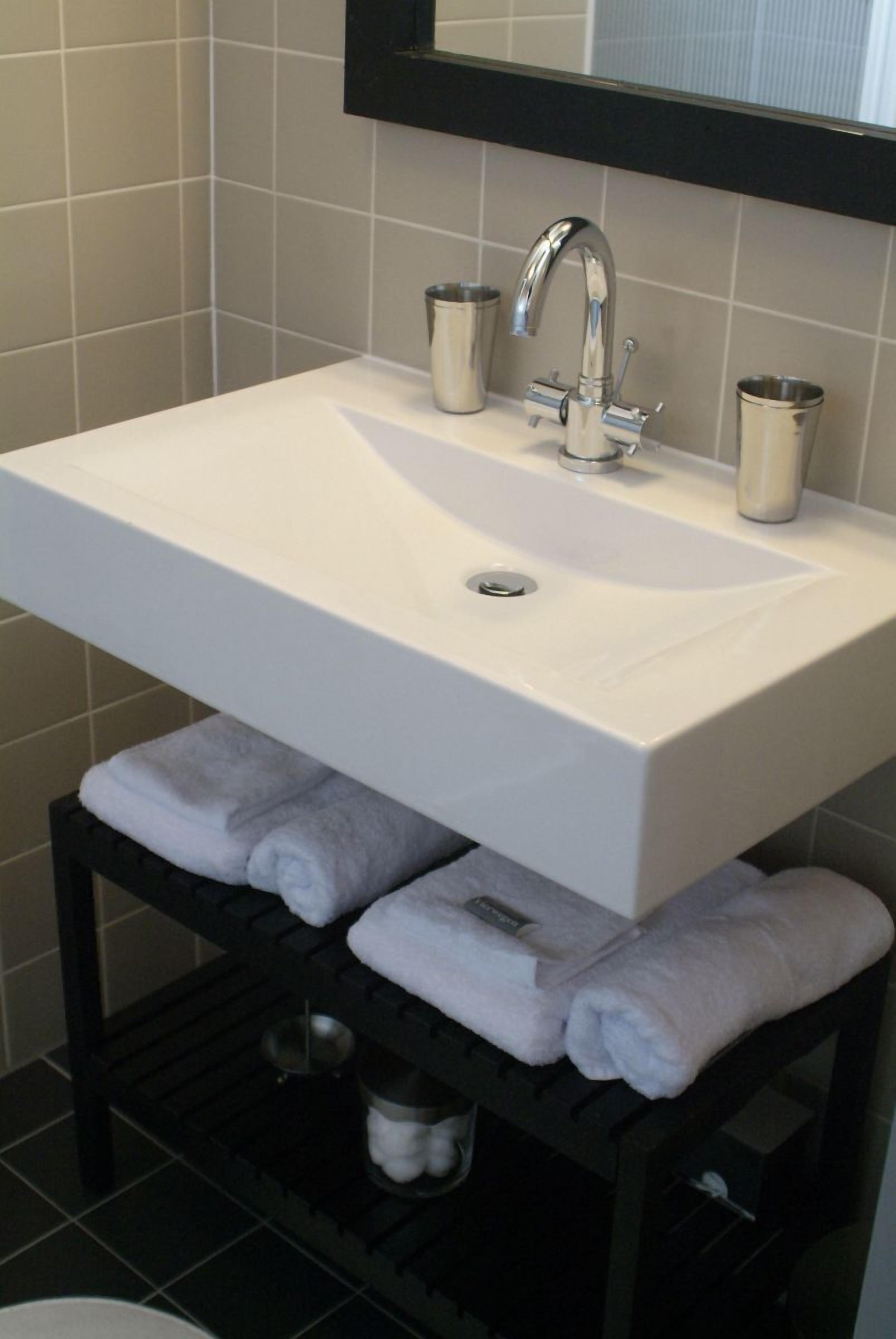 Een badkamer met een wit wastafelmeubel, een spiegel erboven en twee bekers op de rand van de wastafel. Daaronder bevindt zich een handdoekhouder met opgevouwen handdoeken.
