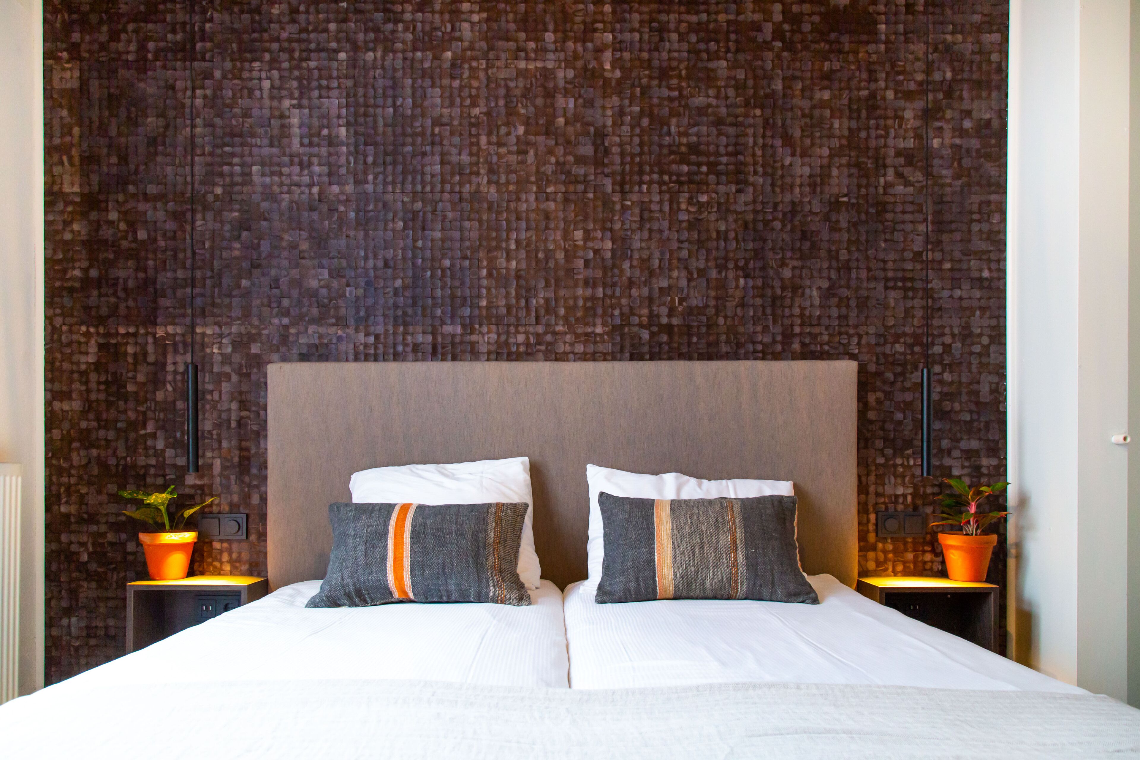 A double bed with two pillows, a brick-patterned wall, and two nightstands with potted plants.