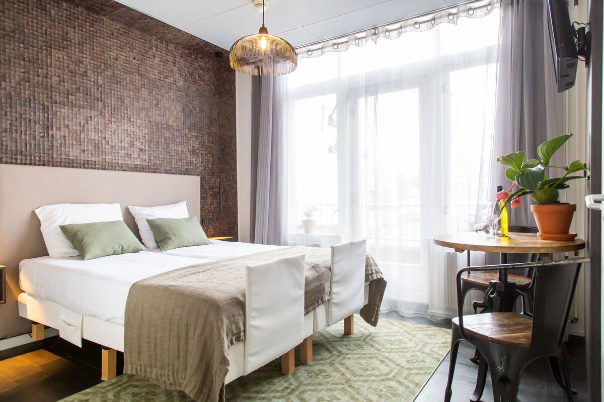 Une chambre avec un mur en briques, un lit avec une literie blanche, un luminaire suspendu, une petite table avec un pot de plante et une chaise.