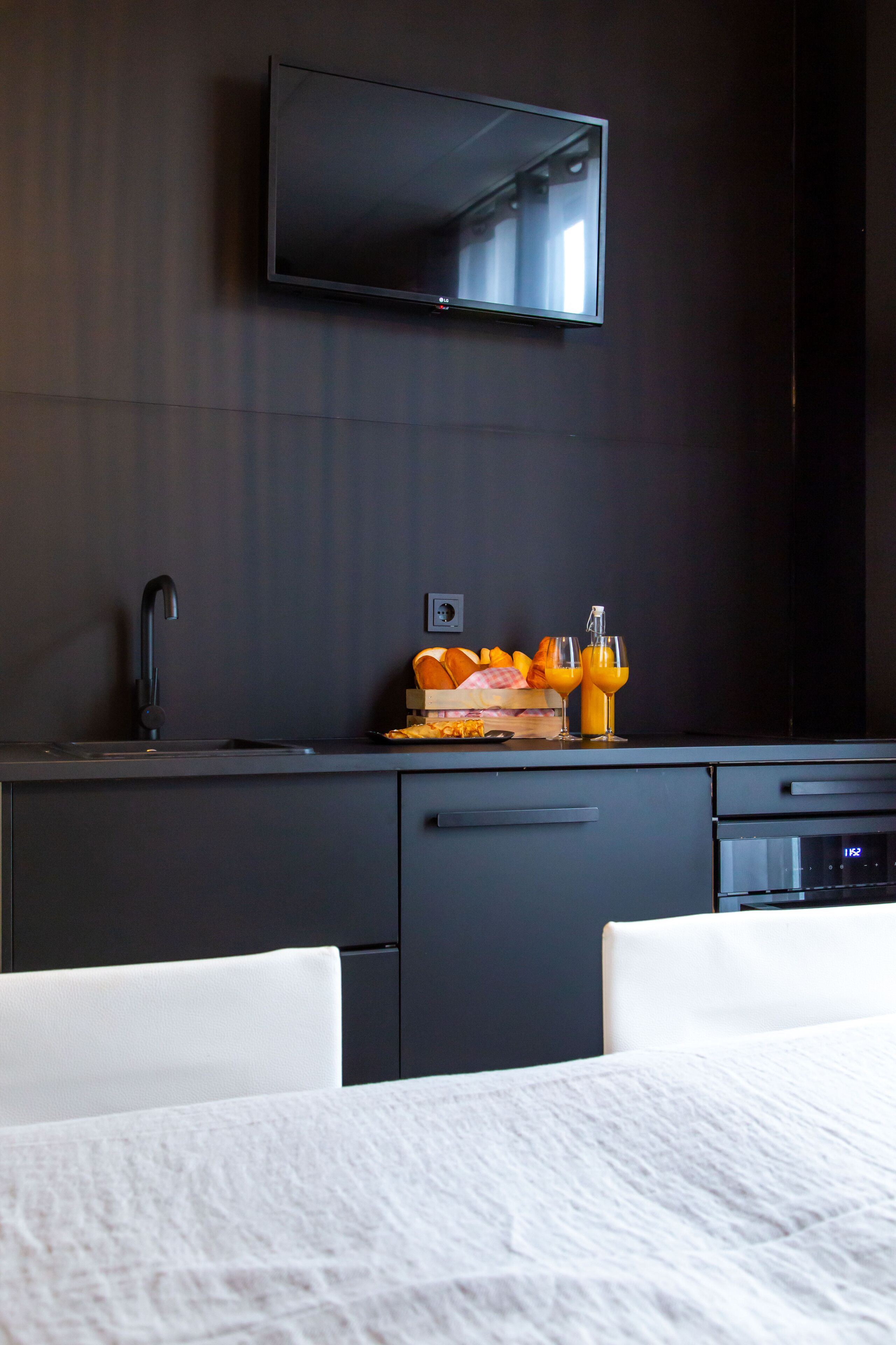 Ein modernes Hotelzimmer mit dunkler Holzvertäfelung, einem flachbildfernseher über einer dunklen Ablagefläche, einem Waschbecken und einer Frühstücksterrasse mit Orangensaft und Brot.