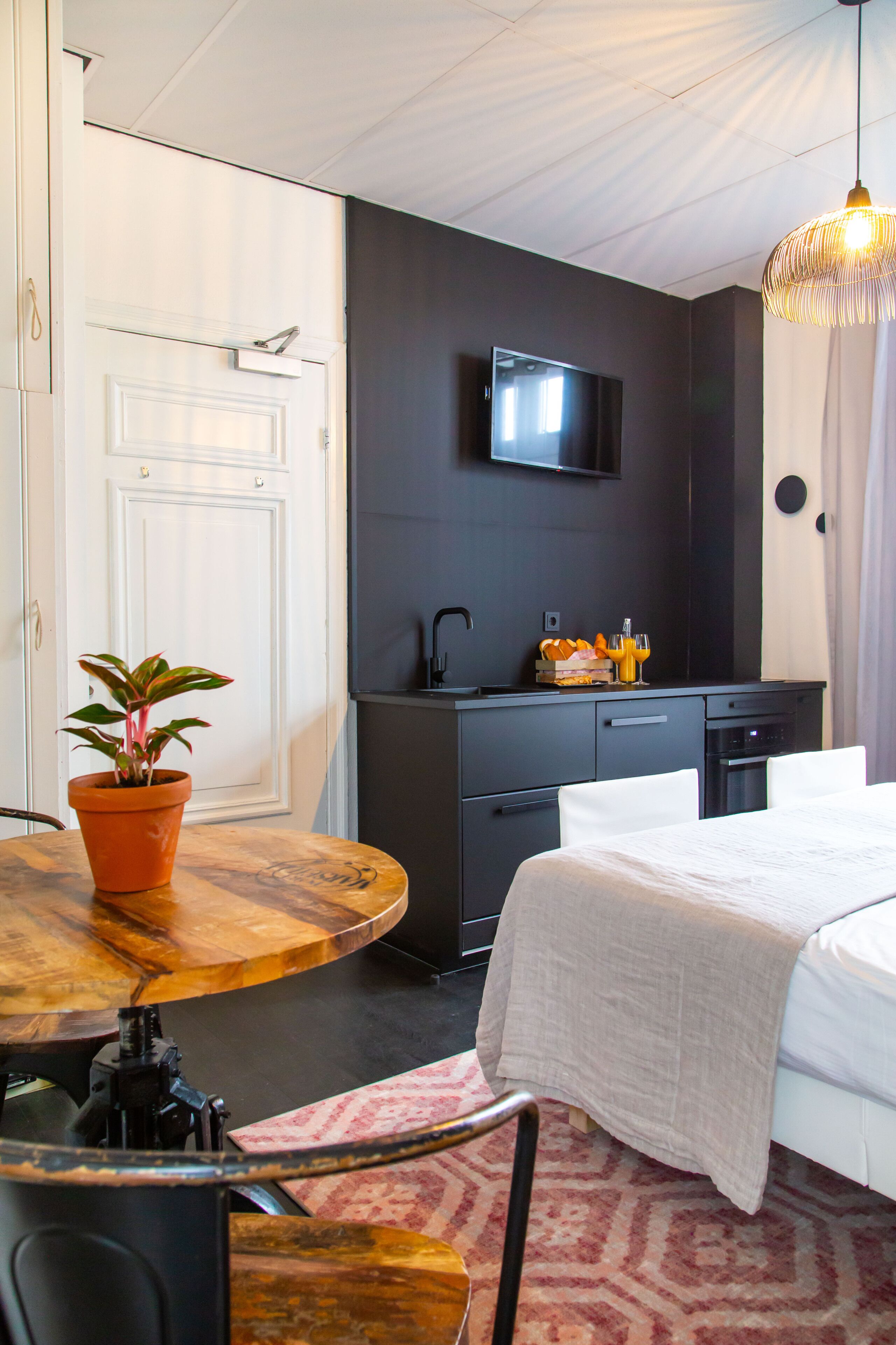 A modern hotel room with a bed, a dining table with a potted plant, a kitchen area with a TV, and a hanging light fixture.