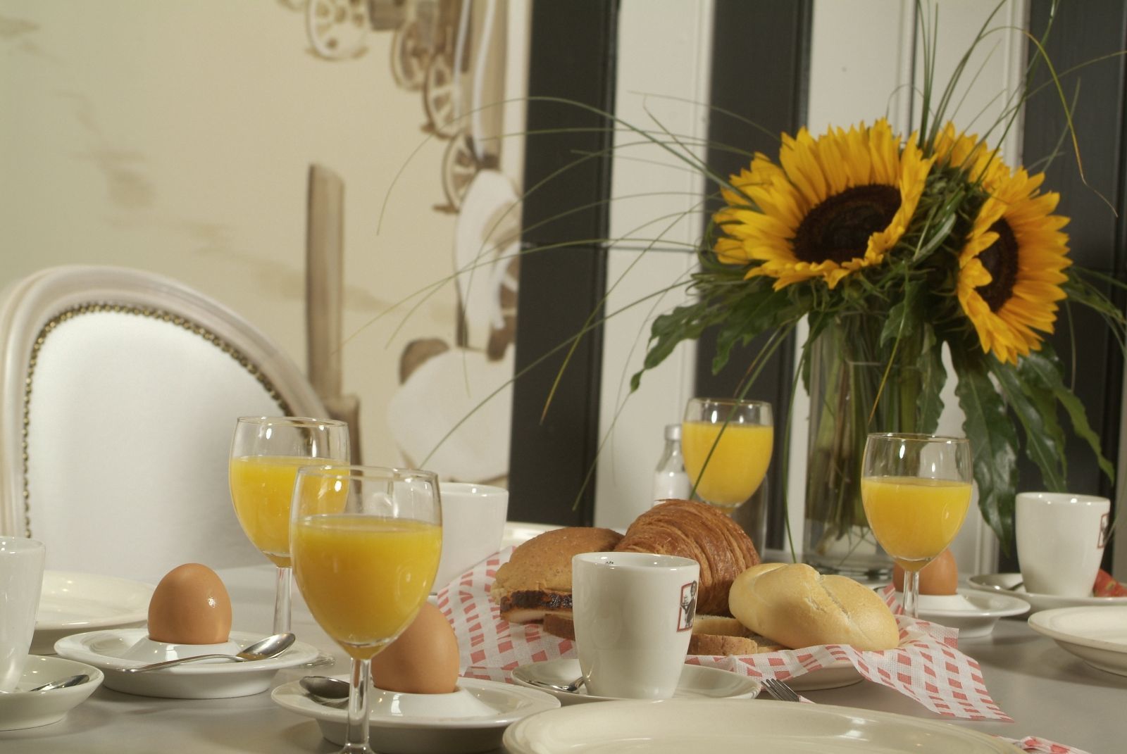 Desayuno con croissants, huevos y jugo de naranja sobre una mesa, junto con un arreglo de girasoles.