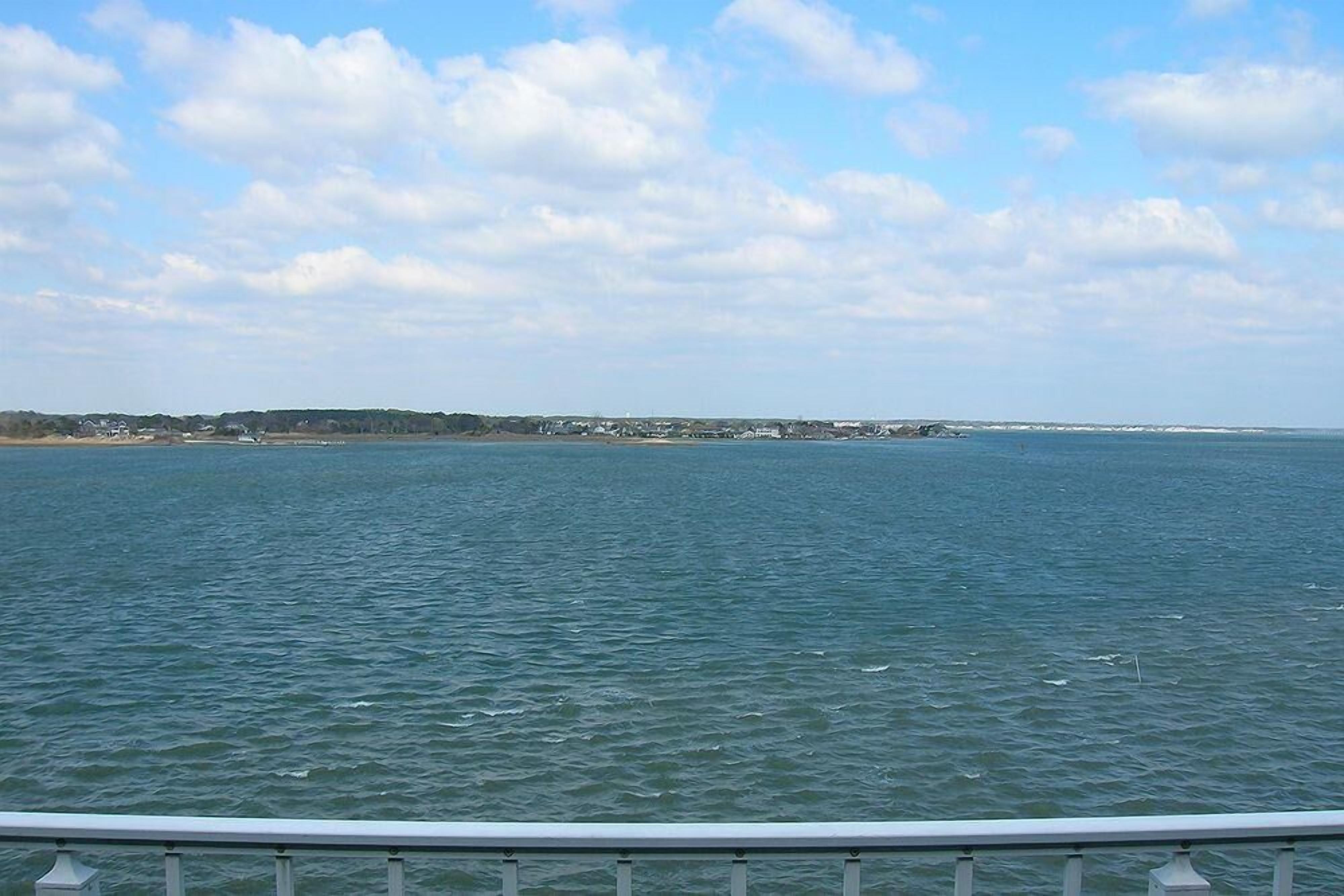 A coastal view from a balcony with a railing, overlooking a calm sea and a distant shoreline with buildings.