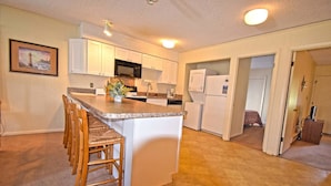 A compact kitchen with white cabinets, a central island with bar stools, a microwave, and a refrigerator. - Severn I 208 3 Br condo by RedAwning (Ocean City)