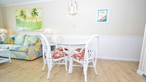 A dining area with a round glass table and white wicker chairs with red cushions. - Shore Lea 102 1 Br condo by RedAwning (Ocean City)