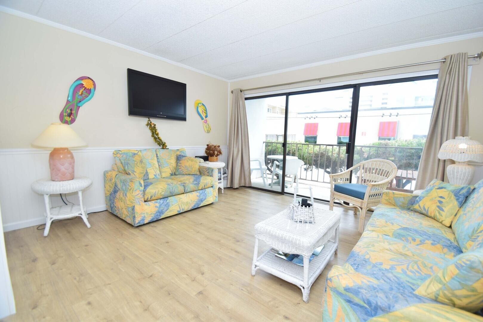 A living room with a sofa, a coffee table, and a television. There is a balcony with outdoor furniture.