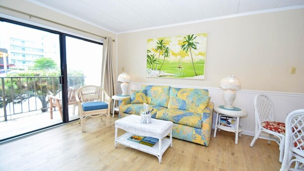 A living room with a sofa, chairs, a coffee table, and a painting of palm trees. - Shore Lea 102 1 Br condo by RedAwning (Ocean City)
