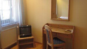 Une petite pièce avec un bureau en bois, une chaise, un téléviseur posé sur un petit meuble et une fenêtre ornée de rideaux légers.