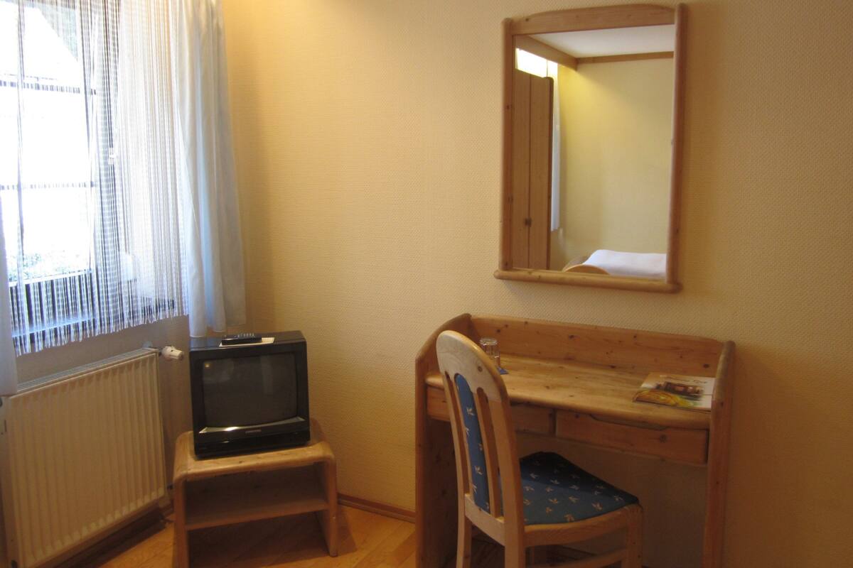 Ein kleines Zimmer mit einem Holztisch, einem Stuhl, einem Fernseher auf einem kleinen Schrank und einem Fenster mit transparenten Vorhängen.