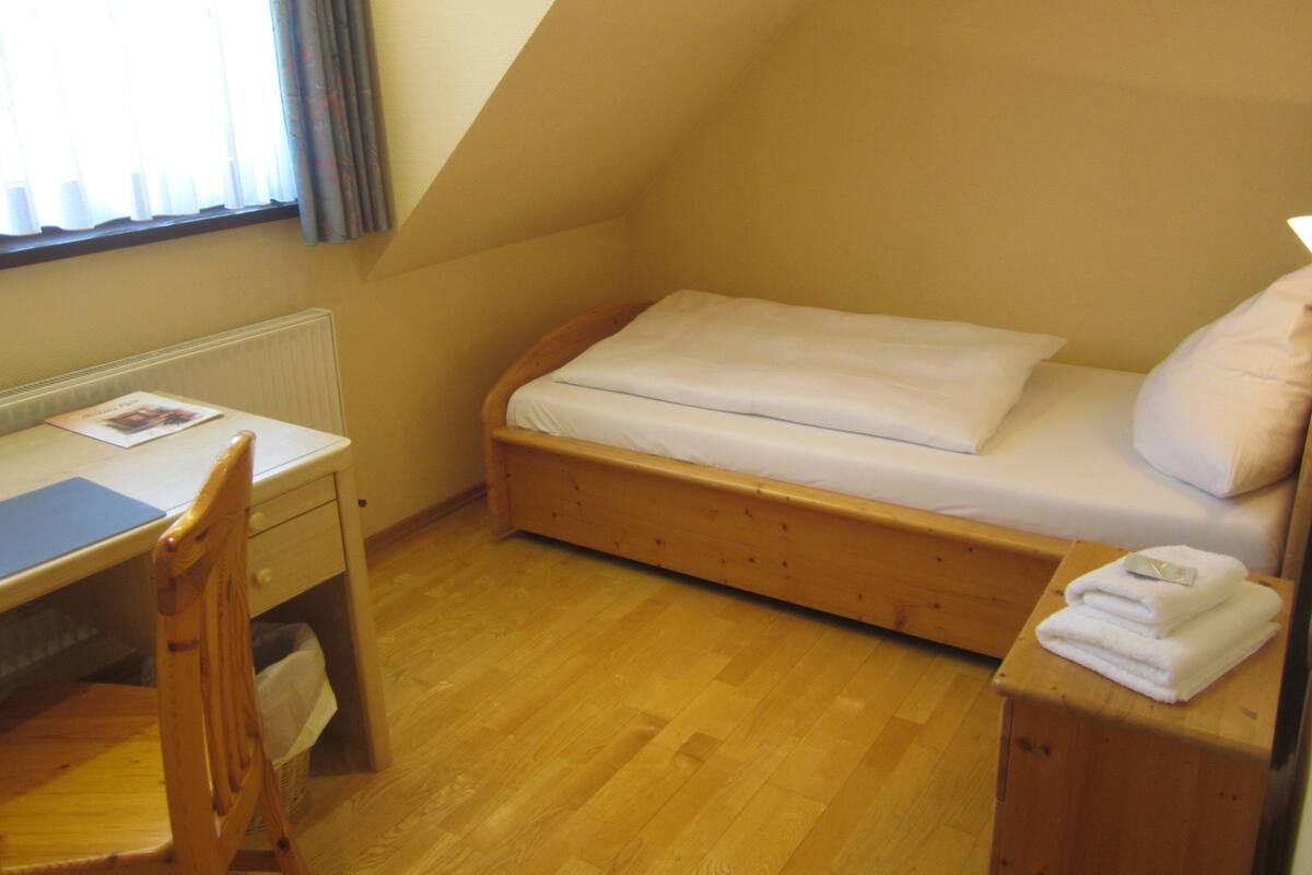 Ein kleines Zimmer mit einem Einzelbett, einem Schreibtisch mit Stuhl und einem Heizkörper.