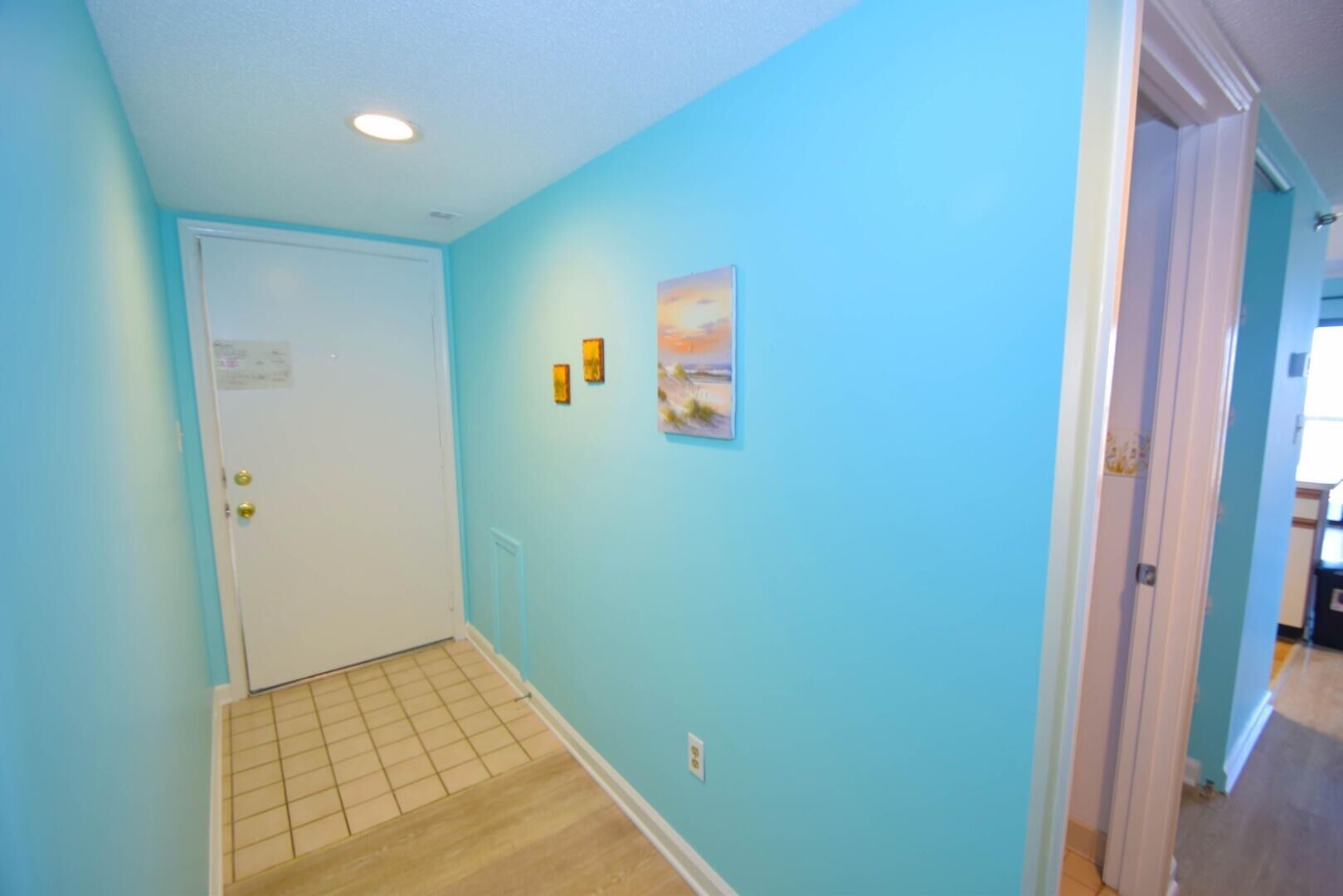 A hallway with light blue walls, a white door, and a tiled floor. There are three small pictures on the wall.