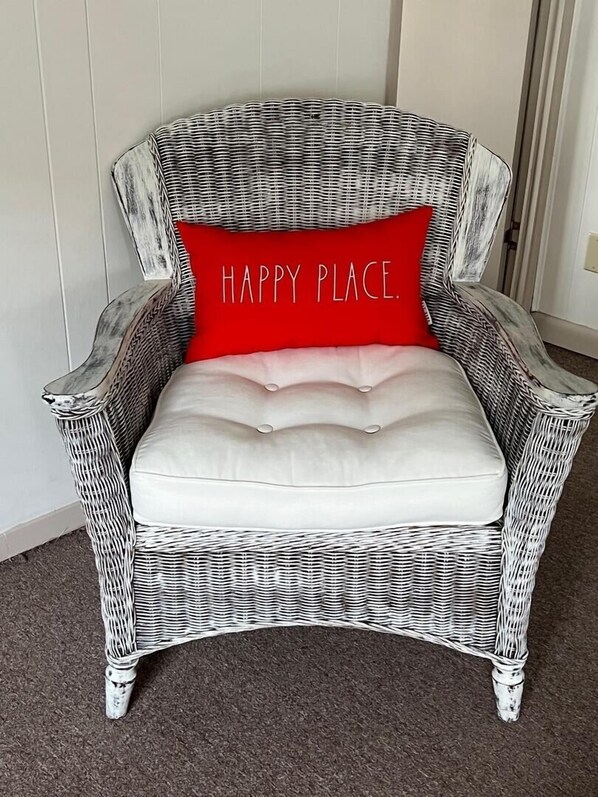 A wicker armchair with a white cushion and a red pillow with the text "HAPPY PLACE."