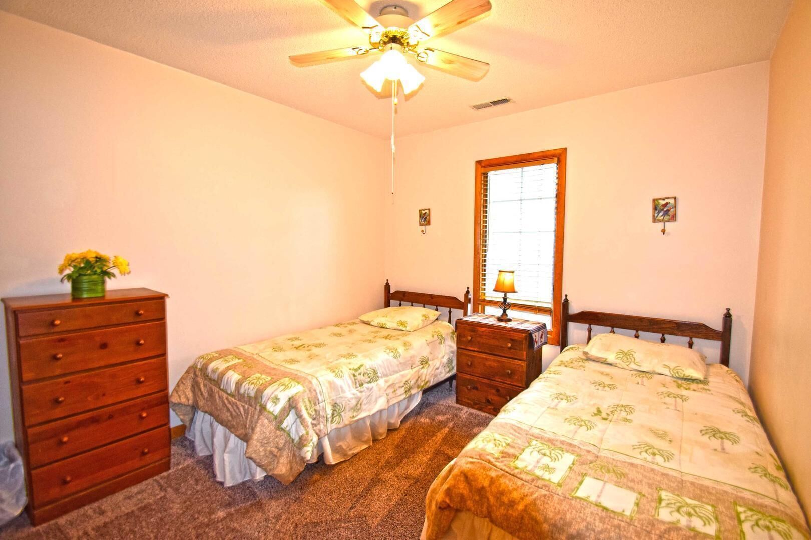 Une chambre avec deux lits simples, une commode en bois, une table de chevet, un ventilateur de plafond et une fenêtre avec des stores.