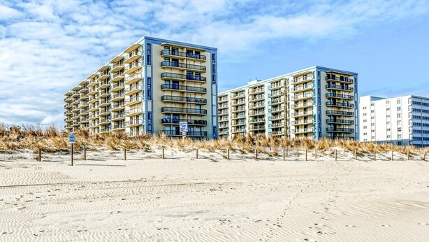 A beachfront hotel with multiple high-rise buildings, a sandy beach in the foreground, and a partly cloudy sky. - Braemar 503N 2 Br condo by RedAwning (Ocean City)