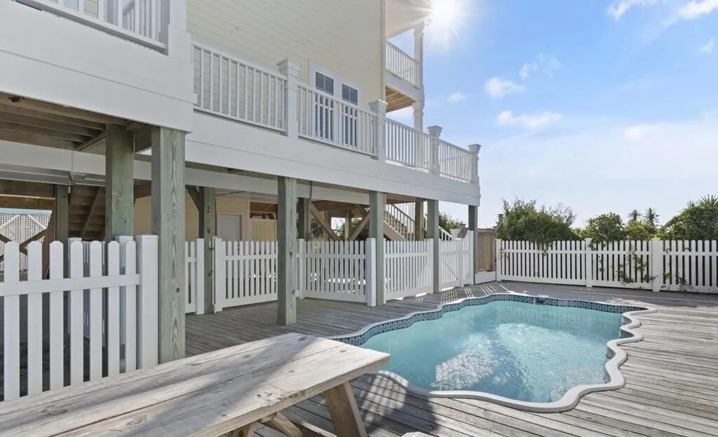 Un bâtiment à deux étages doté d’une terrasse en bois, d’une piscine et d’une clôture blanche en piquets.