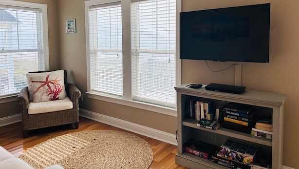 A living room with a flat-screen TV, a bookshelf, a wicker chair, and a large window with blinds.