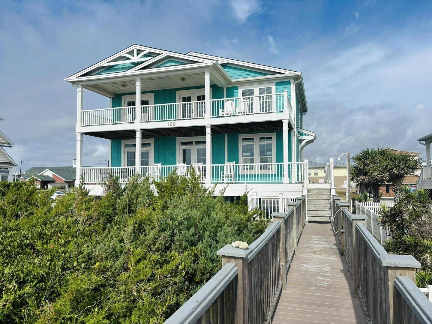 A two-story beachfront property with a wooden deck leading to the entrance.