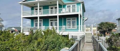 A two-story beachfront property with a wooden deck leading to the entrance.