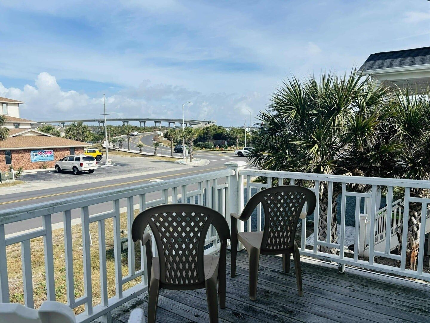 Eine Holzterrasse mit zwei Stühlen, mit Blick auf eine Straße mit Fahrzeugen, ein Gebäude am Strand und eine Brücke in der Ferne.