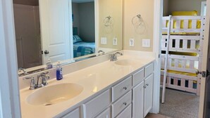 A bathroom with a double sink, a large mirror, and a wooden bunk bed visible through an open door.