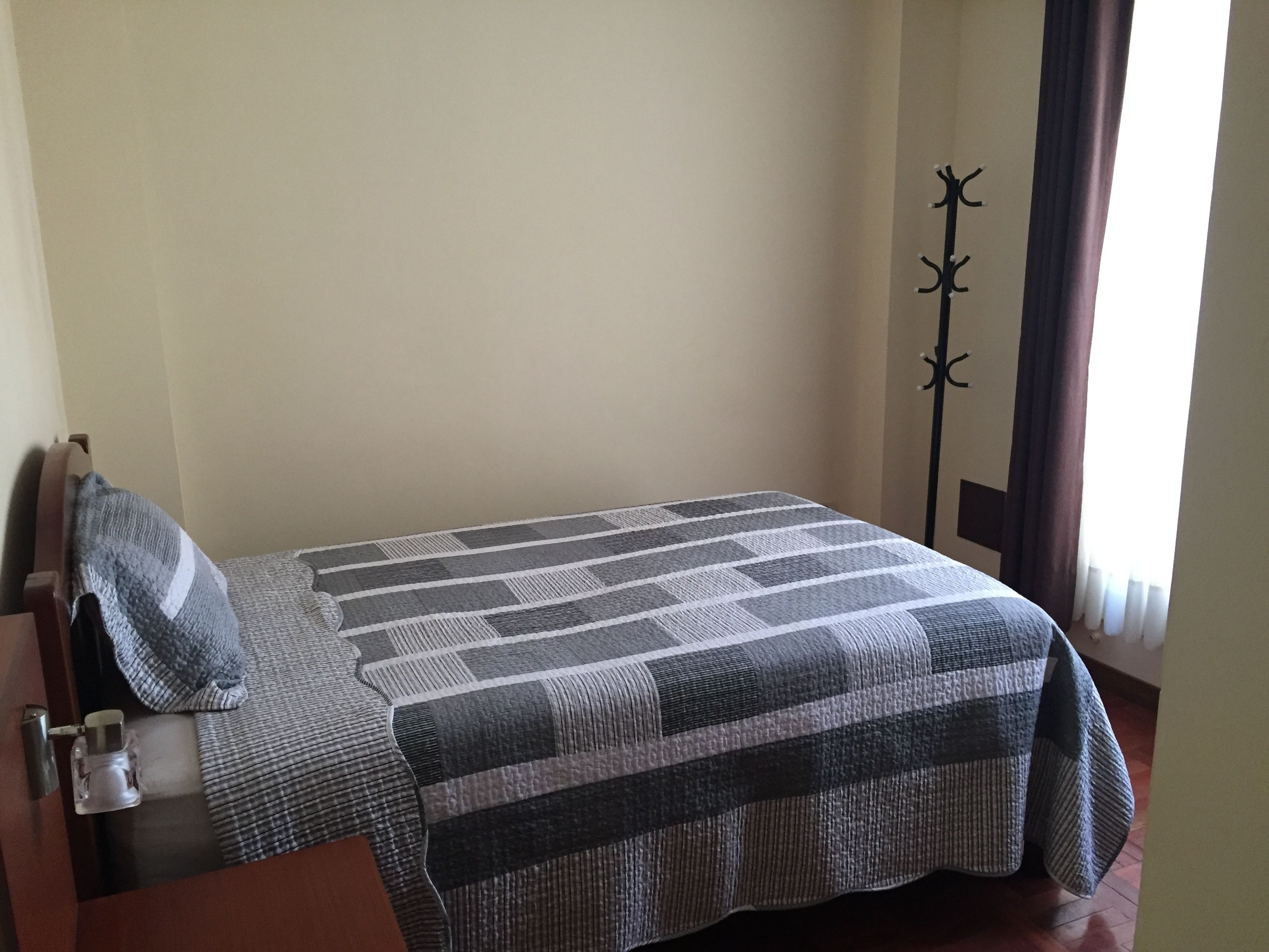 Ein Einzelbett mit gestreifter Decke, ein hölzernes Kopfteil, ein Nachttisch mit Lampe und ein Fenster mit Vorhängen.