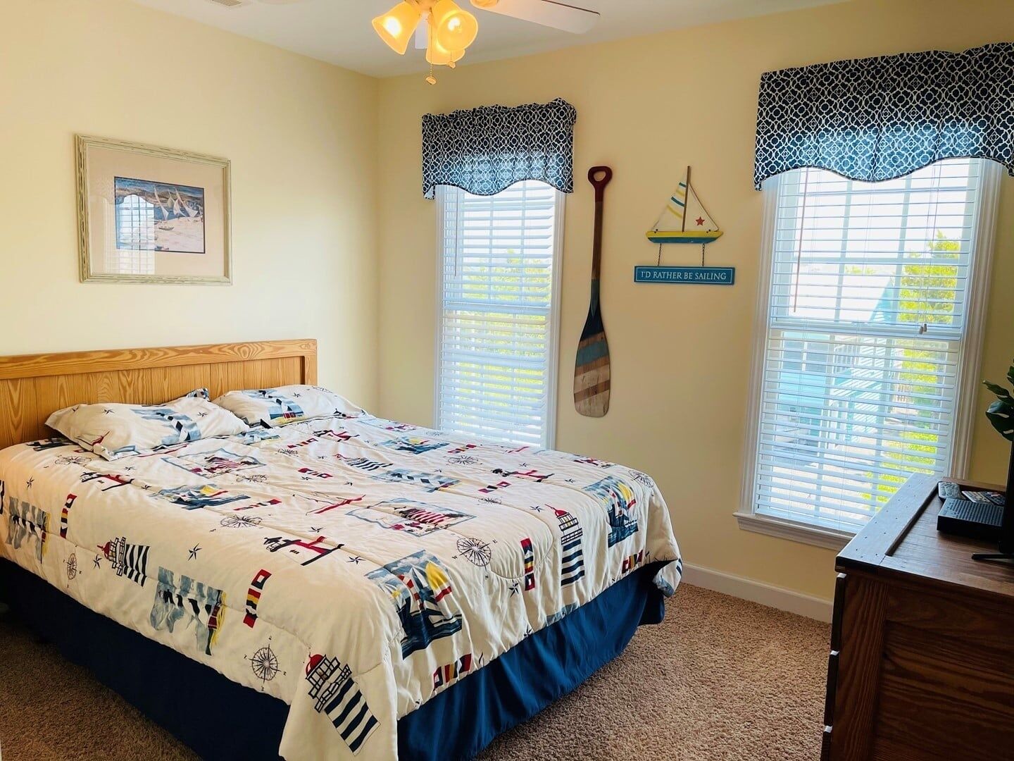 A bedroom with a bed, two windows with patterned curtains, a wall-mounted sailboat decoration, and a framed picture.