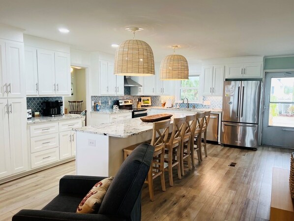 A modern kitchen with white cabinets, a central island, wooden chairs, and a stainless steel refrigerator. - Beach Time 5 Bedroom Home by RedAwning (Supply)