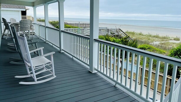 A covered porch with rocking chairs, a view of the beach, and a wooden fence.