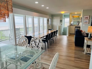 A spacious dining area with a glass table and chairs, a kitchen in the background, and a view of the beach through large windows.
