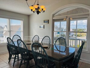 A dining area with a glass-top table, six chairs, a chandelier, and large windows with a view of houses.