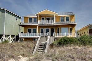 A two-story beachfront house with a balcony, a staircase leading to the entrance, and a sandy foreground. - Carolina Moon 4 Bedroom Home by RedAwning (Supply)