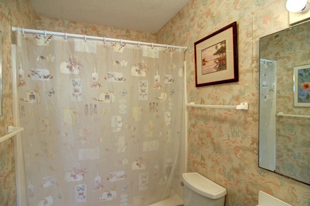 Ein Badezimmer mit Duschvorhang mit floralen und Schmetterlingsmustern, einer Toilette, einem Handtuchhalter und einem Spiegel mit einem gerahmten Bild.